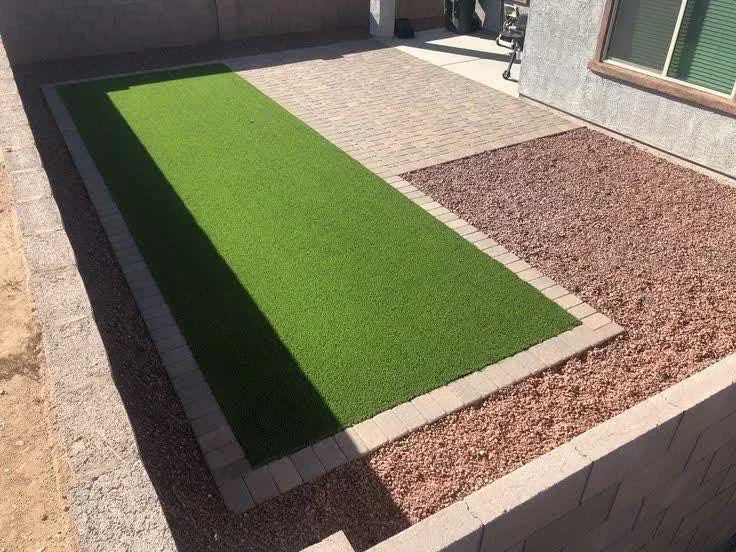 Backyard with bright green synthetic turf strip, tan pavers, and brown gravel sections with concrete edging