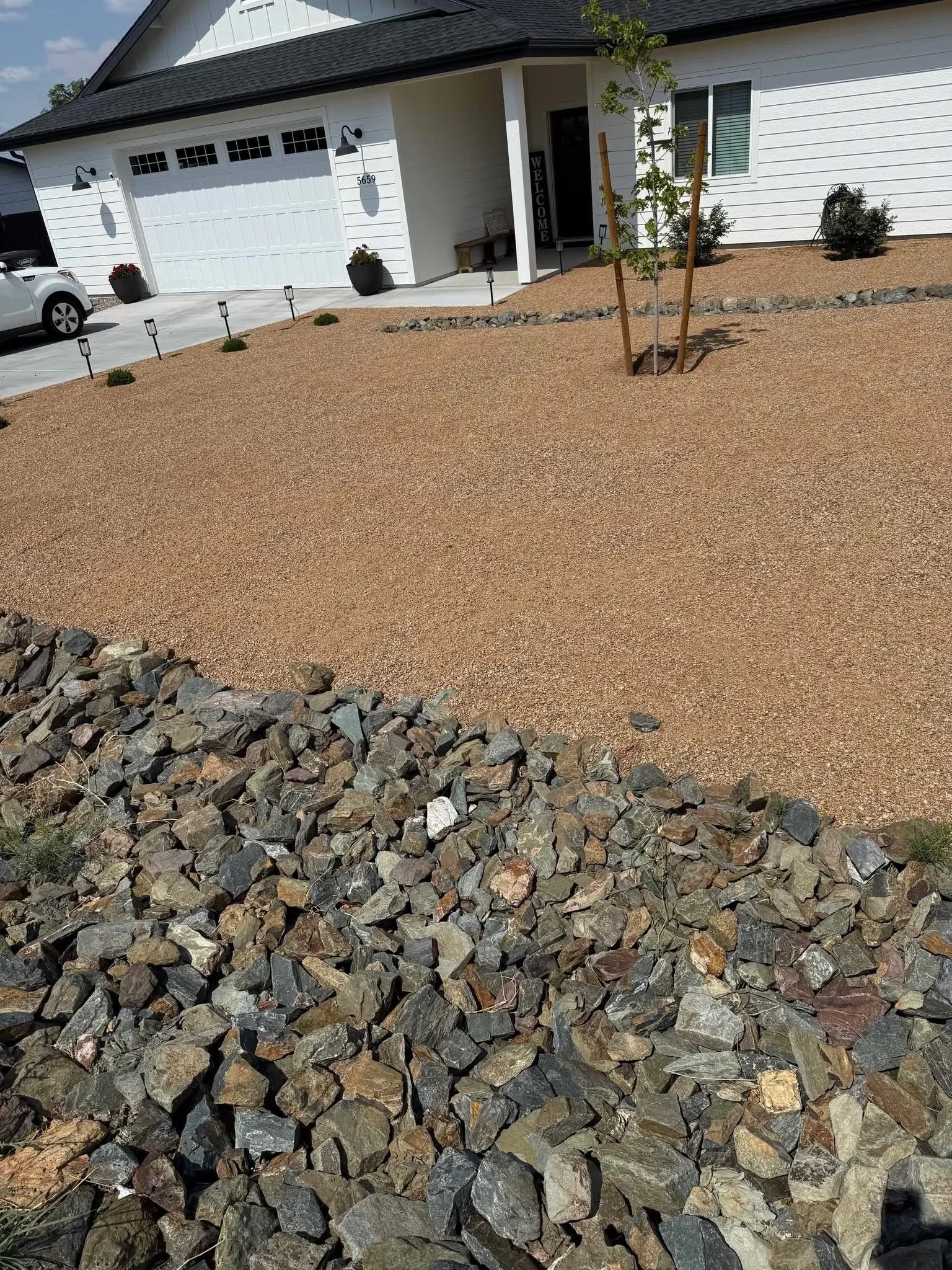 Front yard with tan decomposed granite, dark rock border, young trees, and modern home